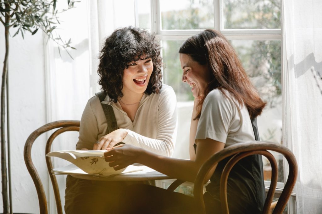 Two women are sitting at a table laughing together whilst reading a newspaper.